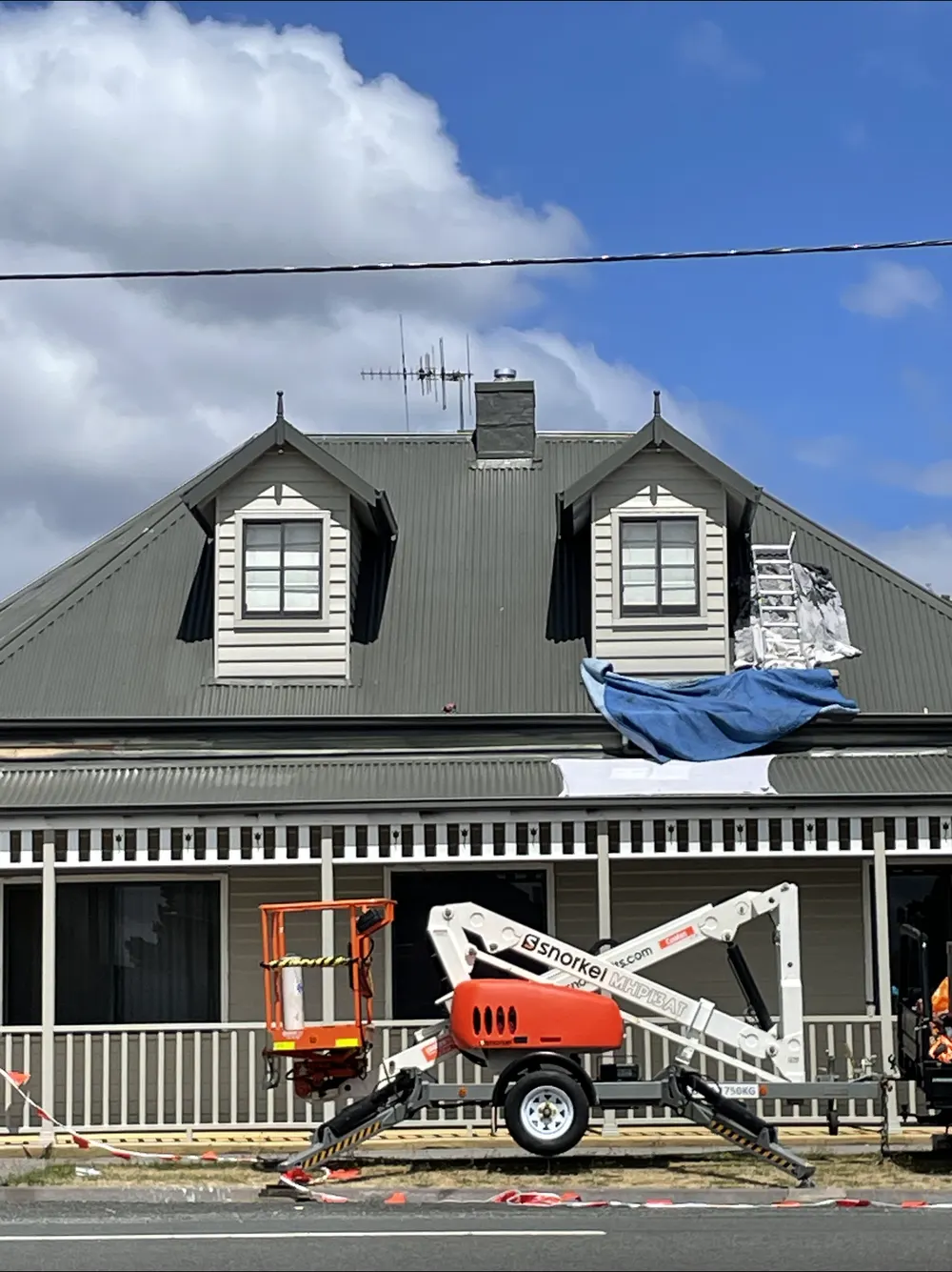 Metal roof with dormer windows, a blue tarp and access equipment in front of the house